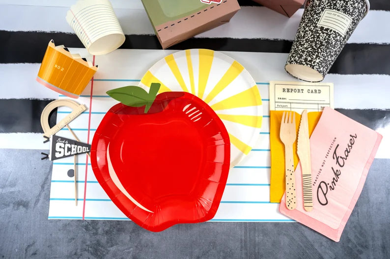 Flat‑lay of back‑to‑school party ware featuring a bright red apple plate on top of a yellow sunburst plate, all arranged on a loose‑leaf paper placemat with assorted classroom‑themed cups, napkins, and utensils.