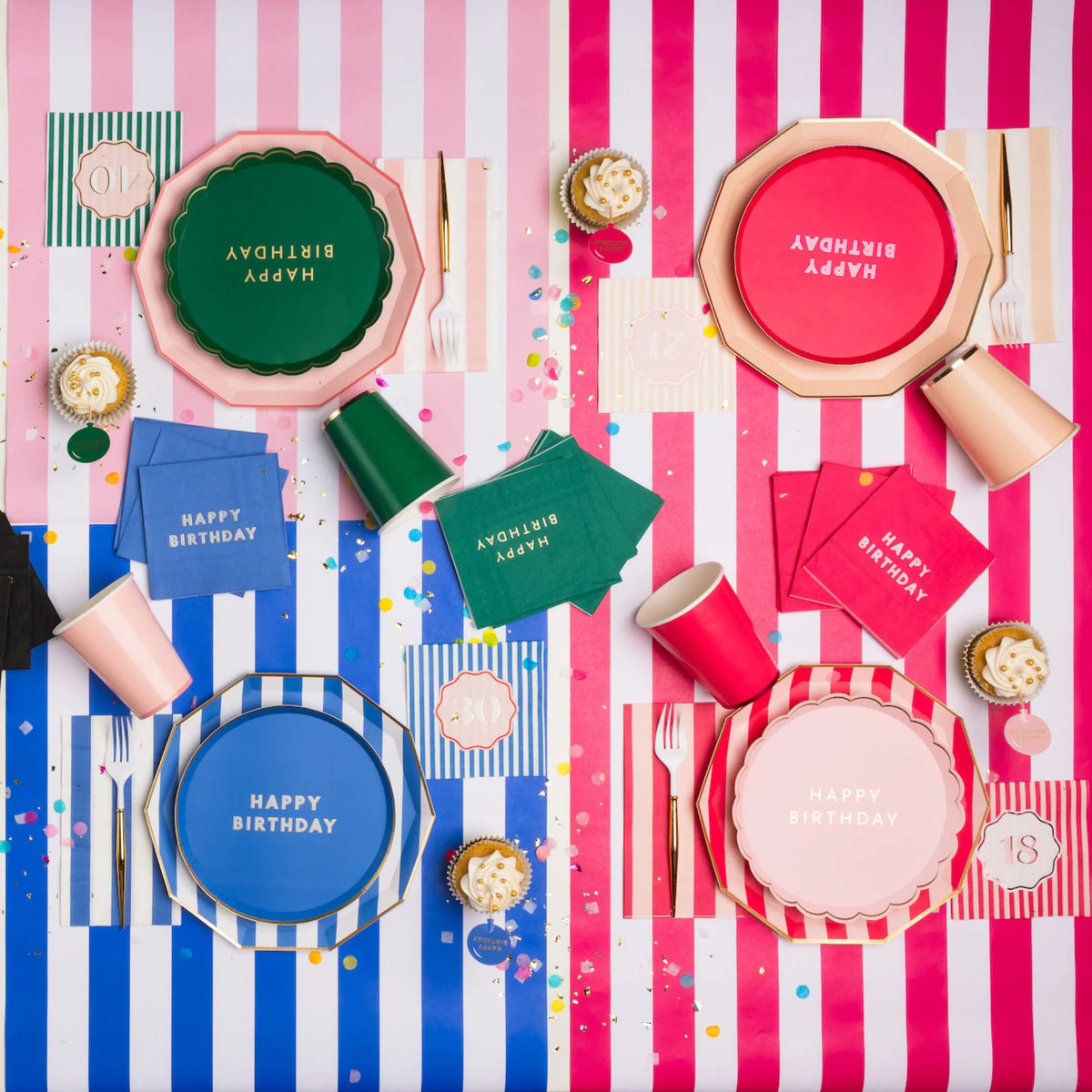Overhead lifestyle shot of colorful milestone birthday party table with Happy Birthday plates in pink blue and green, striped napkins and cups on pink and white striped tablecloths with confetti