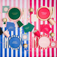 Overhead lifestyle shot of colorful milestone birthday party table with Happy Birthday plates in pink blue and green, striped napkins and cups on pink and white striped tablecloths with confetti