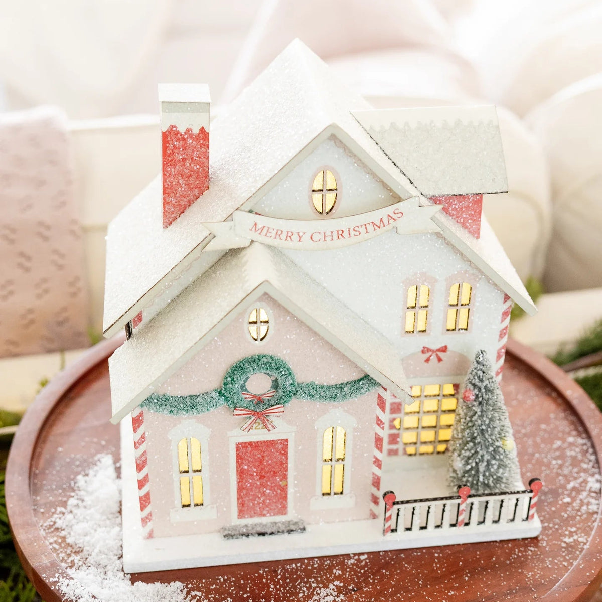 Close-up of the lighted paper house resting on a wood cake stand, showing the warm glow coming from the windows and snow dusting.