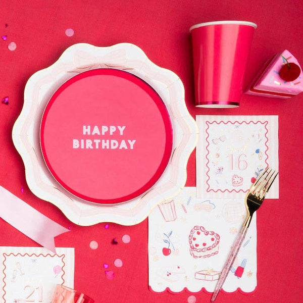 Flat lay of girl's birthday party table with scalloped floral plate, hot pink cup, gold fork, and illustrated birthday napkin on red background