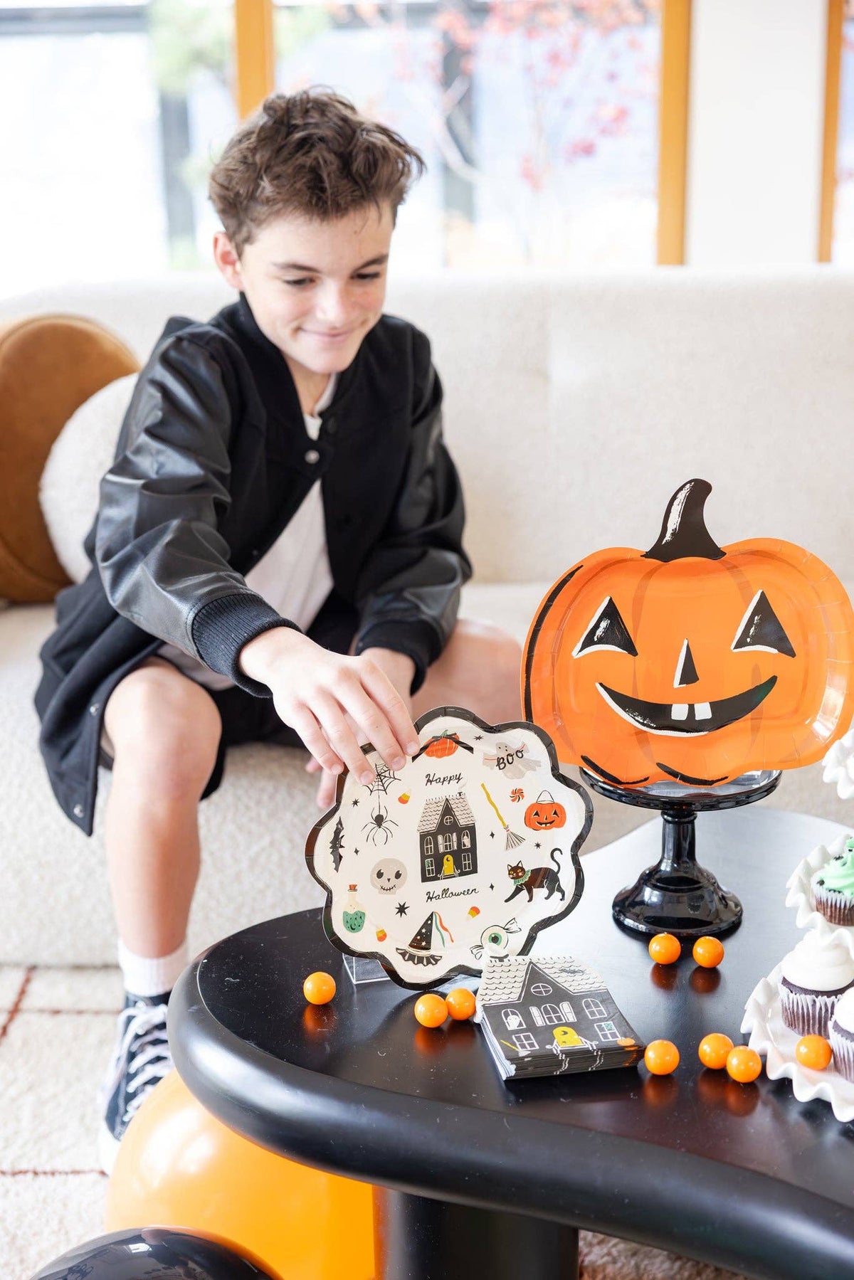 Teen boy in a black bomber jacket reaches for a Happy Halloween scalloped plate on a black coffee table; the orange jack-o-lantern plate sits upright on a black pedestal stand behind it, with a stack of haunted-house napkins and orange gumballs nearby. Close-up angled lifestyle shot.