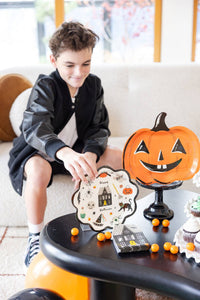 Teen boy in a black bomber jacket reaches for a Happy Halloween scalloped plate on a black coffee table; the orange jack-o-lantern plate sits upright on a black pedestal stand behind it, with a stack of haunted-house napkins and orange gumballs nearby. Close-up angled lifestyle shot.