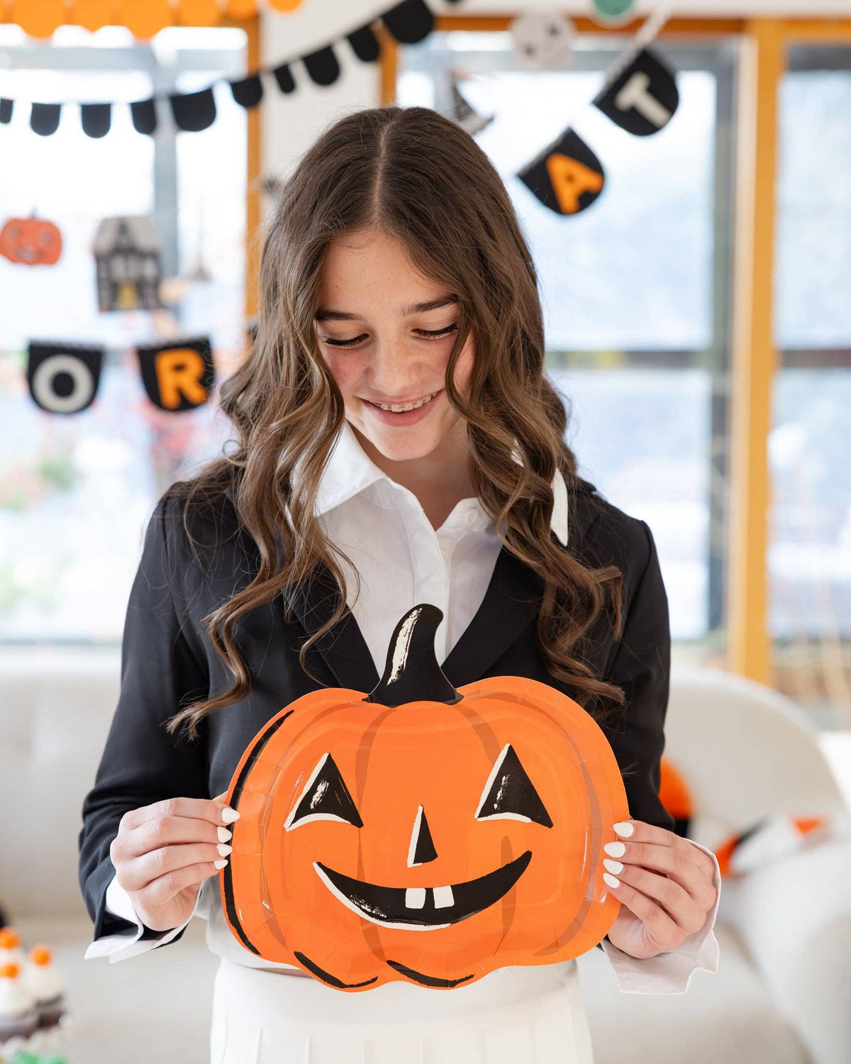 Teen girl looking down and smiling at the jack-o-lantern plate she holds in both hands; soft-focus Halloween banners ("OR," "TREAT," scalloped, icon garland) fill the background. Angled lifestyle portrait.