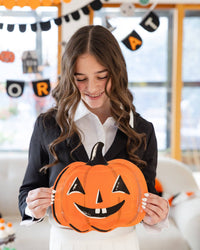 Teen girl looking down and smiling at the jack-o-lantern plate she holds in both hands; soft-focus Halloween banners ("OR," "TREAT," scalloped, icon garland) fill the background. Angled lifestyle portrait.