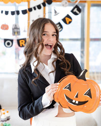 Smiling teen girl in a black blazer and white collared shirt holds the orange jack-o-lantern paper plate at her waist, mouth open mid-laugh; behind her, layered Halloween banners including an orange-letter "TREAT" circle banner, a black scalloped banner, and an icon garland. Angled lifestyle shot, bright window light.
