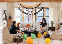 Wide living-room dessert-table scene: a black "TRICK OR TREAT" circle-letter banner with an orange-and-black scalloped garland and a Halloween icon string above; the jack-o-lantern plate stands on a black pedestal beside a Happy Halloween scalloped plate, haunted-house favor boxes, cupcakes, and a "Stay Spooky" pennant; orange, green, yellow, and black balloons scattered on the rug. Straight-on environmental shot.