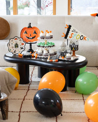 Straight-on dessert table: jack-o-lantern plate on a black stand, Happy Halloween scalloped plate, a "Stay Spooky" multicolor pennant flag, black-and-white haunted-house favor boxes, "Happy Spooky Season" napkins, and tiered chocolate cupcakes; balloons in the foreground.