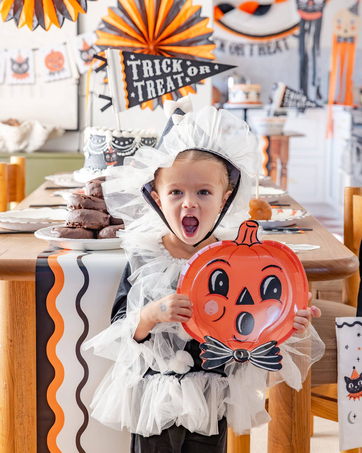 Child in a Halloween costume holding a pumpkin plate both with surprised face at a Halloween decorated table.