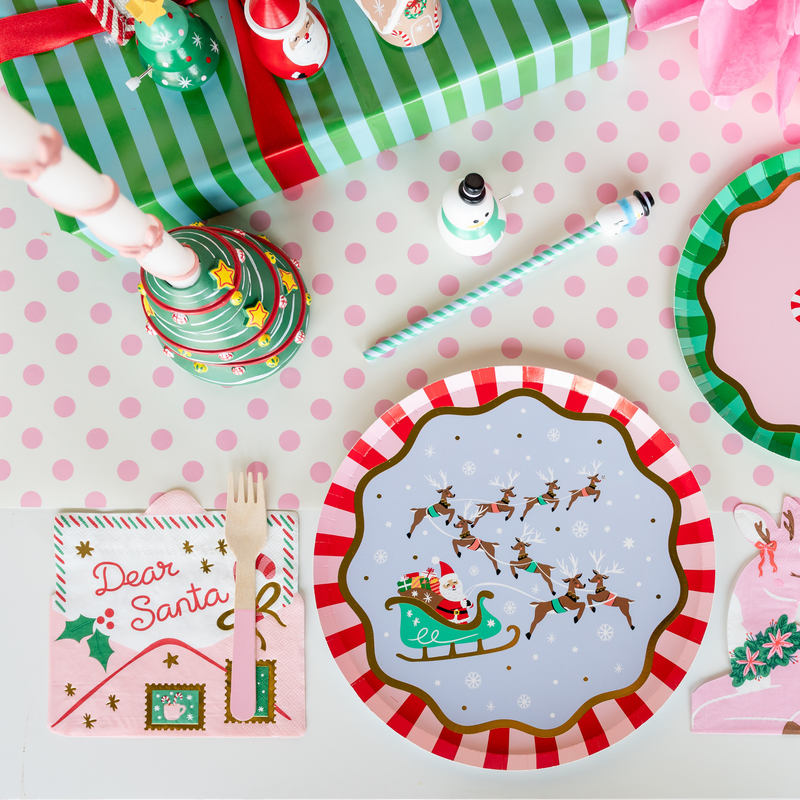 Overhead kids' holiday place setting showing the "Dear Santa" napkin with the Santa sleigh plate, a green plate, pink flatware, and Christmas décor on a pink polka-dot runner.