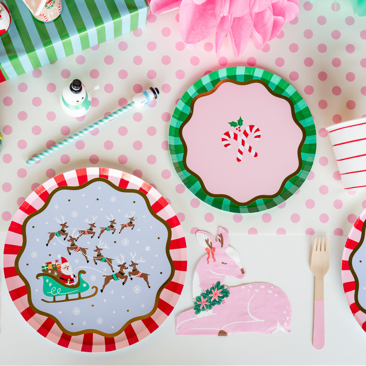 Overhead kids' holiday place setting showing the Santa sleigh plate with a green dessert plate, a candy cane plate, pink flatware, and Christmas décor on a pink polka-dot runner.