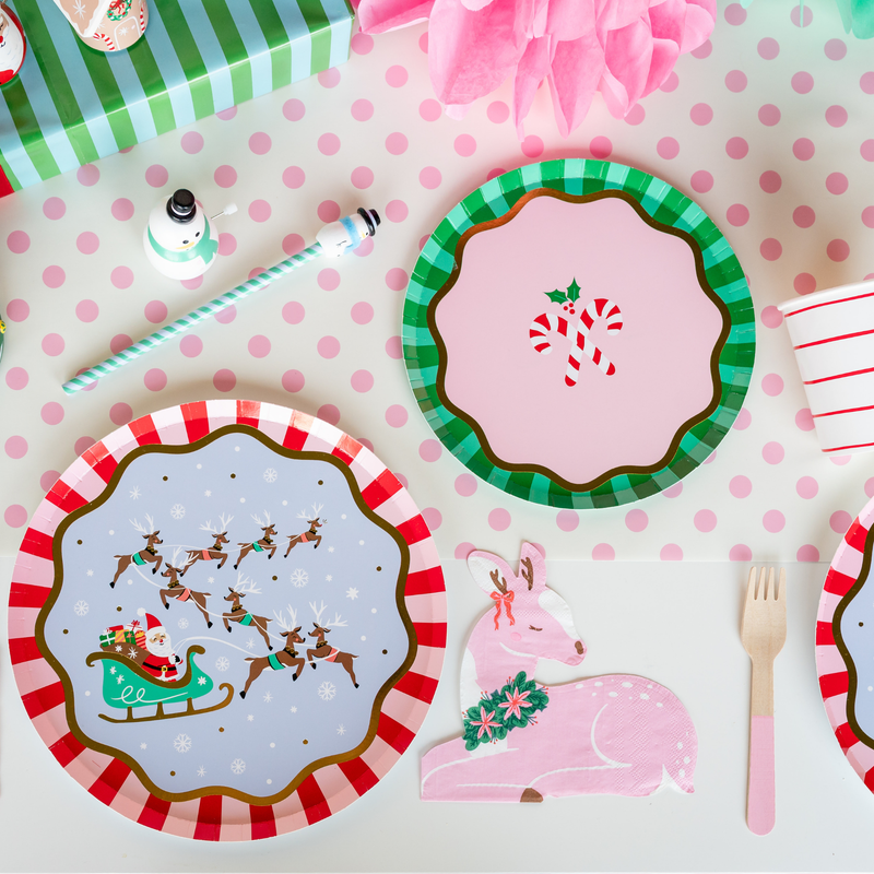 Overhead kids' holiday place setting showing the Santa sleigh plate with a green dessert plate, a candy cane plate, pink flatware, and Christmas décor on a pink polka-dot runner.