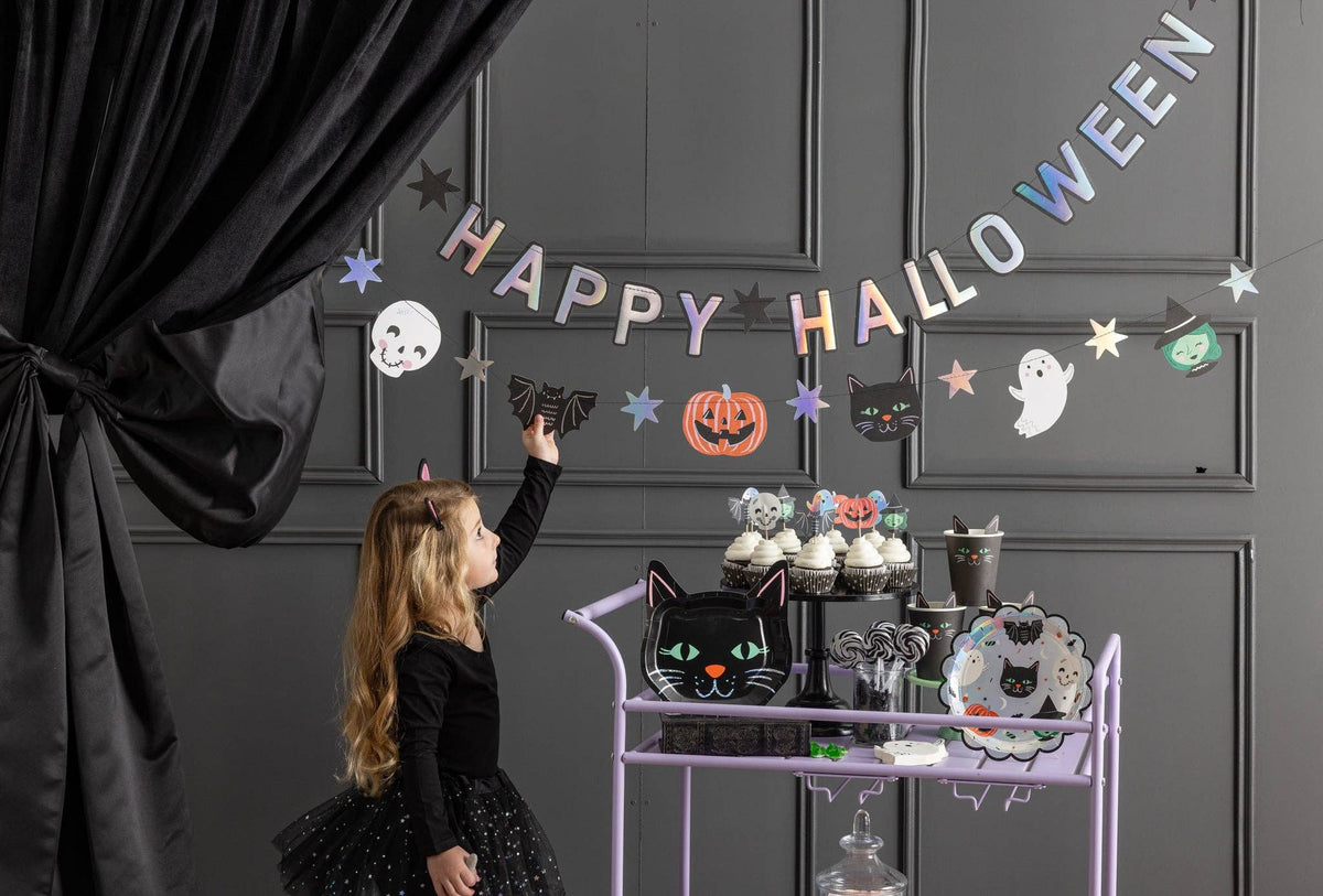 Styled kids' Halloween party scene showing the banner above a purple rolling cart loaded with treats, cupcakes, and spooky decorations, with a woman and child.