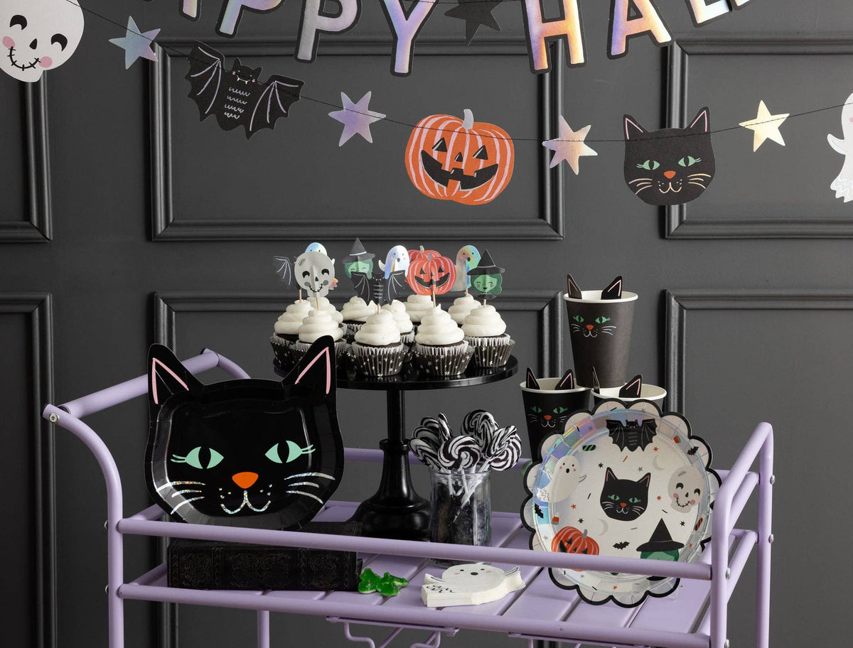 Wide kids' Halloween party scene showing the SPO collection — purple rolling treat cart, cat plates, cupcakes with character toppers, black cups, and the holographic "Happy Halloween" banner on a dark gray paneled wall.
