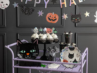 Wide kids' Halloween party scene showing the SPO collection — purple rolling treat cart, cat plates, cupcakes with character toppers, black cups, and the holographic "Happy Halloween" banner on a dark gray paneled wall.