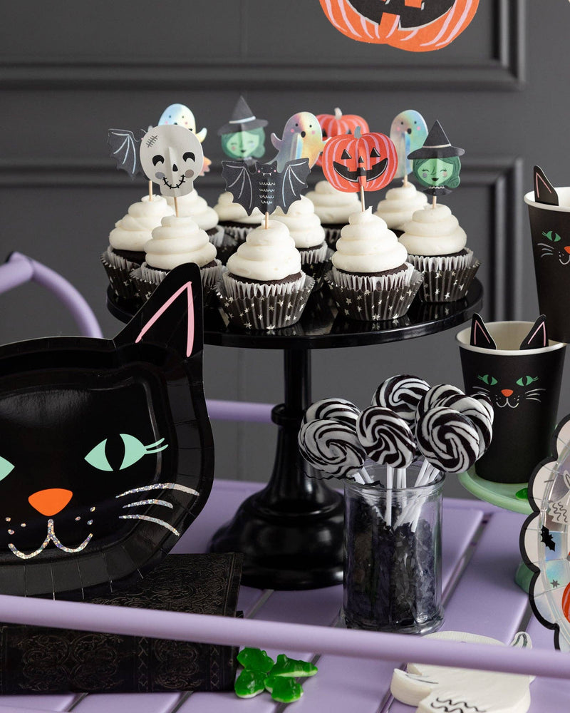 Styled Halloween dessert table featuring frosted cupcakes decorated with Spooky Friends paper toppers on a black cake stand, next to a shaped black cat paper plate.
