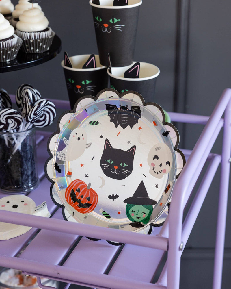 Styled Halloween party cart in light purple, displaying the Spooky Friends scalloped paper plates alongside matching black cat cups and frosted cupcakes.