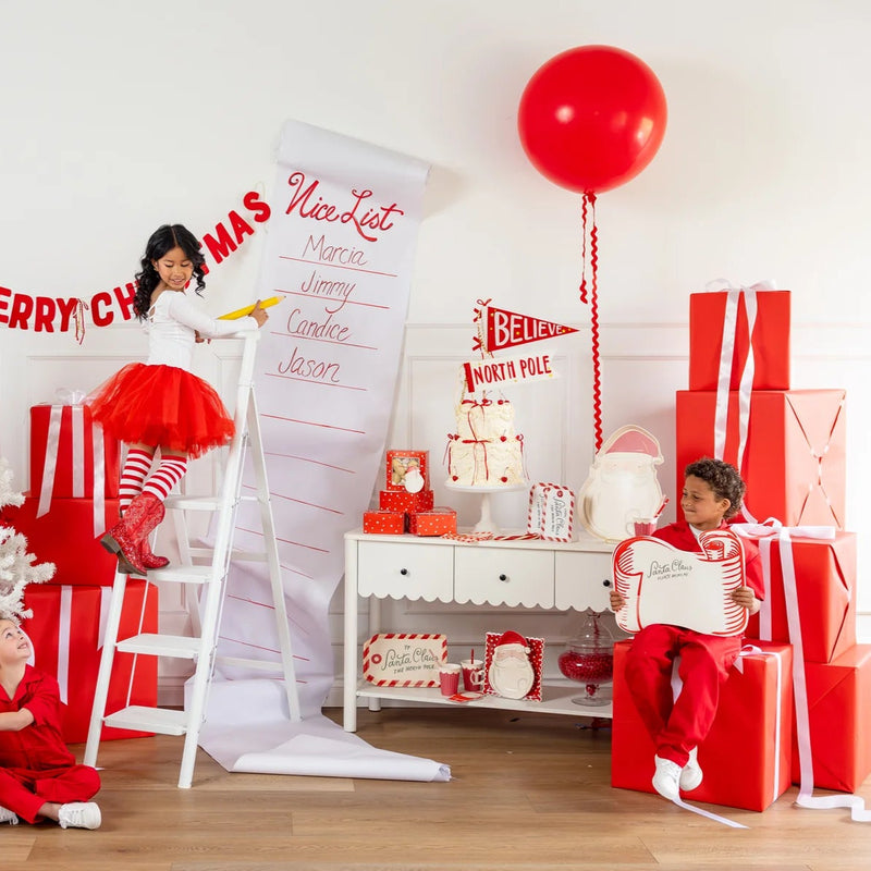 Two children playing next to a giant Nice List scroll and a boy holding the large Santa placemats in a festive room.