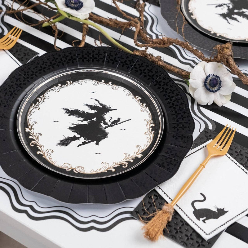 Layered Halloween place setting showing a vintage witch paper plate resting on a black die-cut charger, paired with a gold fork and black cat cocktail napkin.