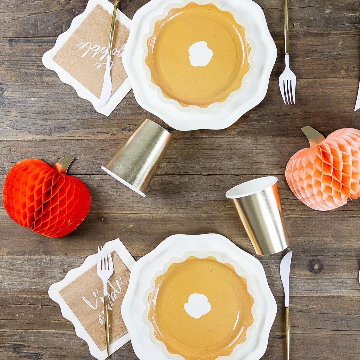 Overhead Thanksgiving table with pumpkin décor, gold cups, and kraft “Le gobble” cocktail napkins by pie plates.