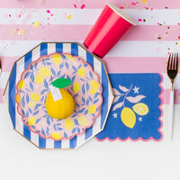 Lemon‑themed place setting with layered blue stripes, citrus plates, cobalt lemon napkin, and gold flatware.
