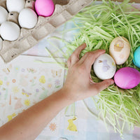 A lifestyle shot of a hand applying a temporary bunny tattoo to a white egg, surrounded by colorful eggs and spring party supplies.