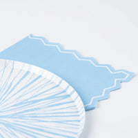 Close-up of the upper right corner showing the die-cut zigzag edge with white inset border line against a white surface. A blue-and-white sunburst paper plate is partially visible at lower left, sitting on top of the stacked napkins.