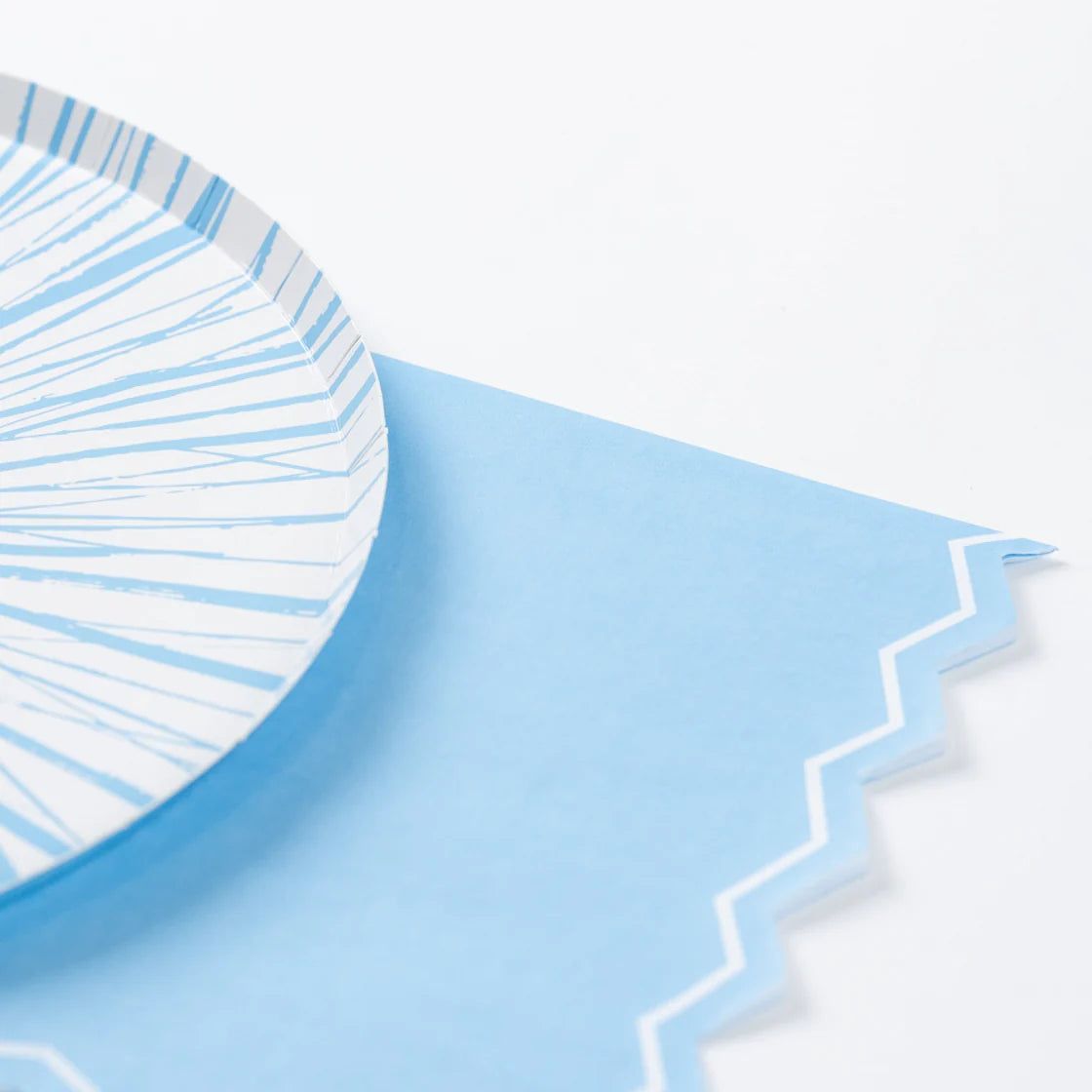 Close-up of the right side and lower corner of the napkin showing the angular zigzag die-cut edge with white border line. A blue-and-white sunburst paper plate sits partially on top of the napkin at left, its blue rim edge and radiating brushstroke lines visible.