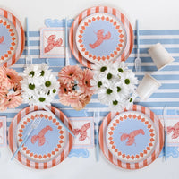 Overhead of a table set for four to six on a blue-and-white stripe table runner. Light blue zigzag napkins visible at each place setting positioned under or beside lobster cocktail napkins. Lobster print salad plates over orange stripe charger plates at every setting. Blue forks, peach gerberas and white daisies as centerpieces.