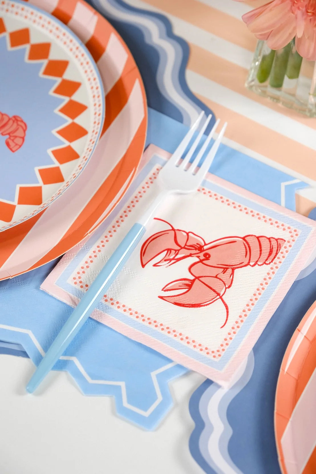 Close-up of the light blue zigzag napkin positioned to the right of a lobster place setting. The lobster cocktail napkin rests on top of the zigzag guest towel. White fork with blue handle lies across both. The zigzag edge is clearly visible against the blue scalloped placemat border. Peach stripe runner and gerbera daisies visible in background.