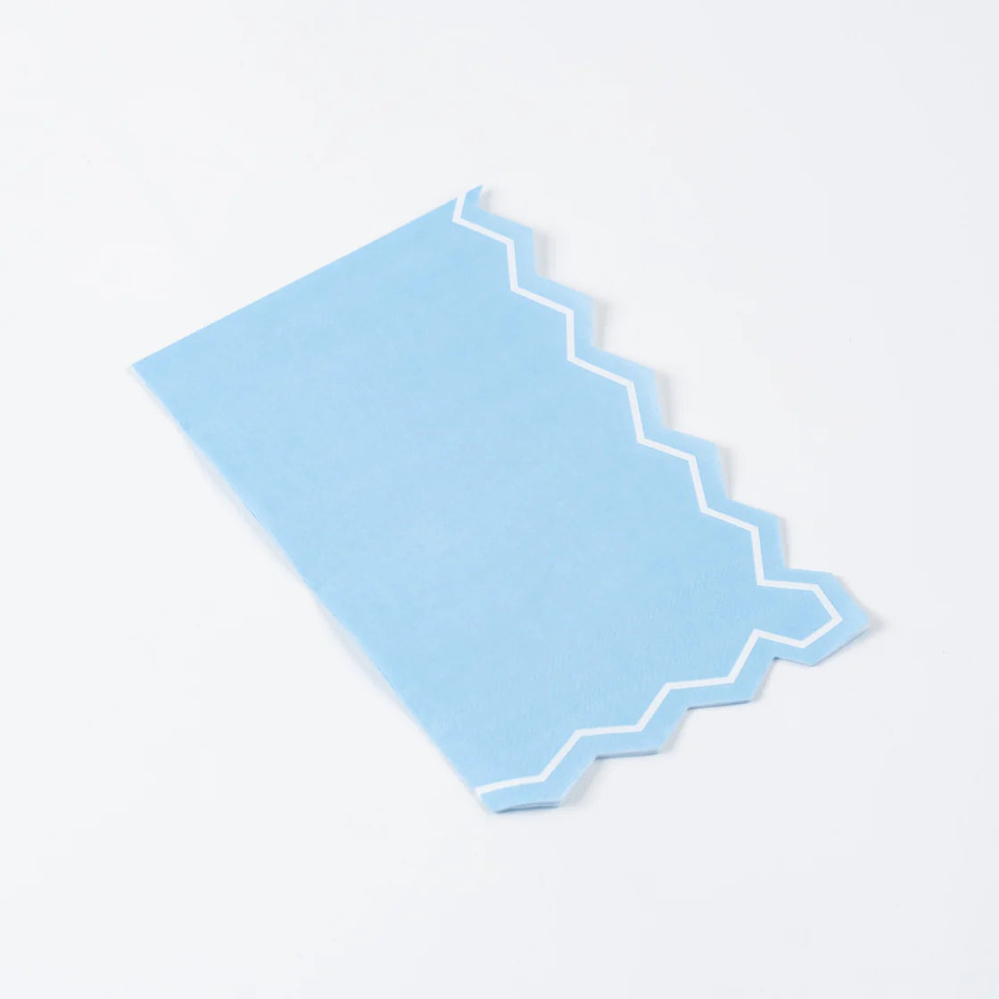 Single light blue zigzag-edge paper guest towel on a white background, angled diagonally. Full shape visible: rectangular portrait orientation with straight left edge and top, die-cut zigzag on right edge and bottom. White inset border line runs parallel to the zigzag cut. Matte paper texture visible.