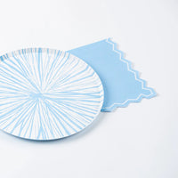 The full blue-and-white sunburst paper plate resting on top of the light blue zigzag-edge napkin against a white surface. The zigzag border is visible protruding from under the plate at the upper right and lower right. The plate features light blue radiating brushstroke lines from a central point on a white ground.