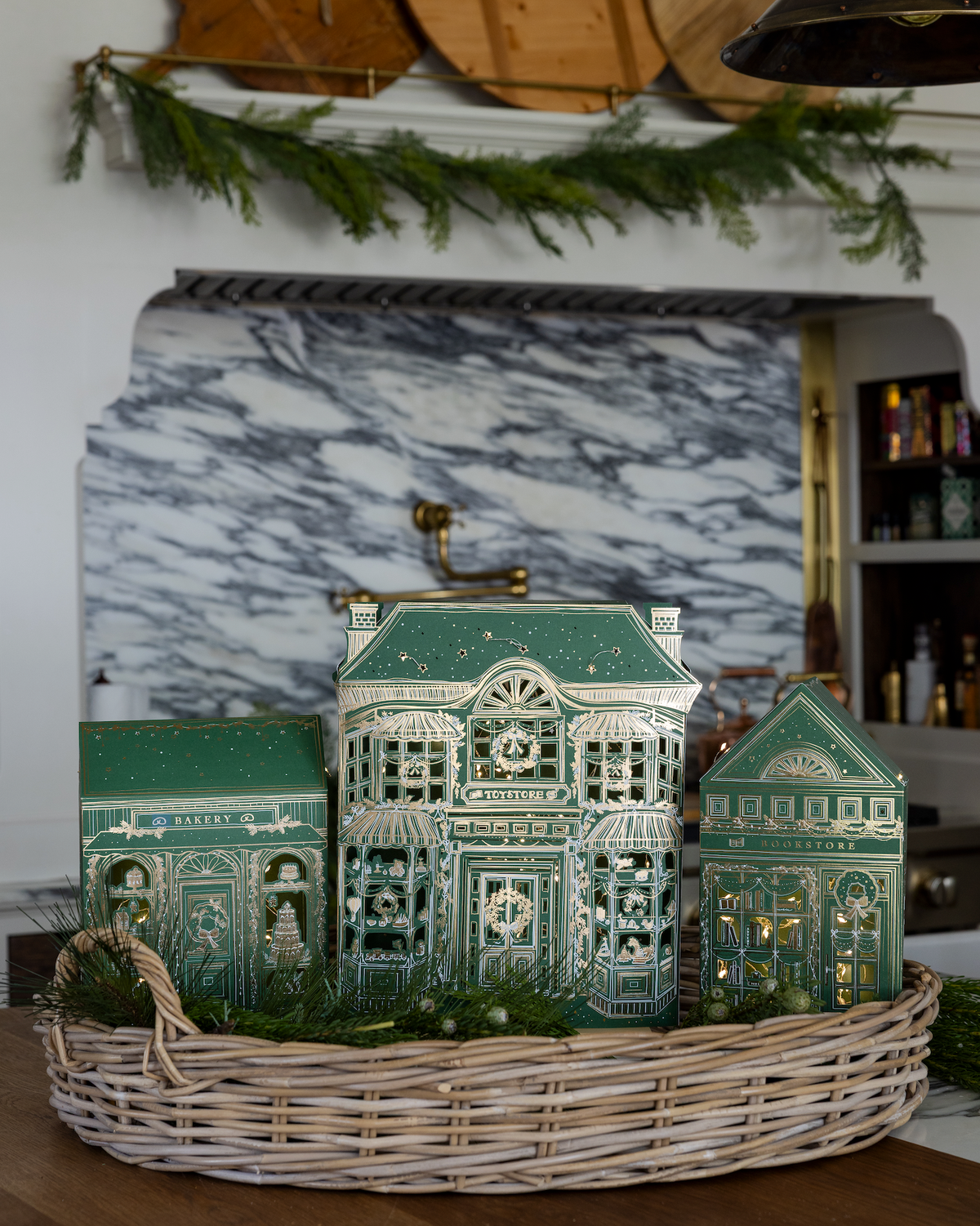 Three lit paper Christmas village shops nestled in a wicker basket with greenery on a kitchen counter in front of a marble backsplash