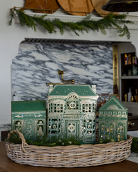 Three lit paper Christmas village shops nestled in a wicker basket with greenery on a kitchen counter in front of a marble backsplash