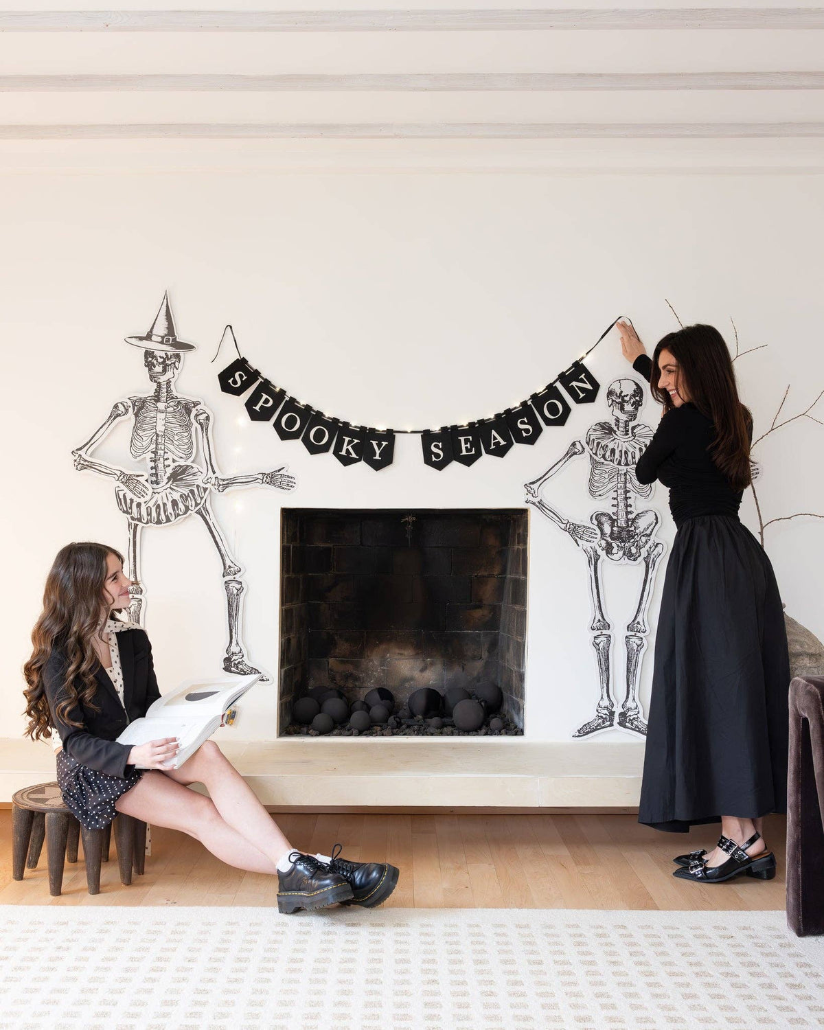 Two women decorating a fireplace with skeleton and 'Spooky Season' banner for Halloween.