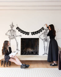 Two women decorating a fireplace with skeleton and 'Spooky Season' banner for Halloween.