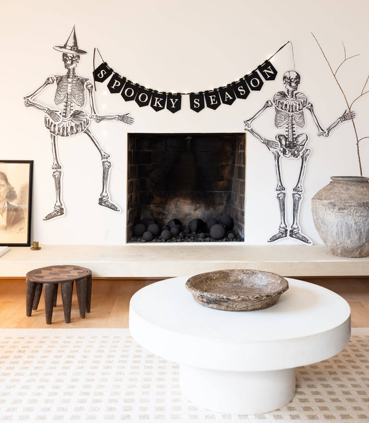 Living room with Halloween decorations including skeletons and a 'Spooky Season' banner hung across a mantel in a room, fairy lights illuminated.