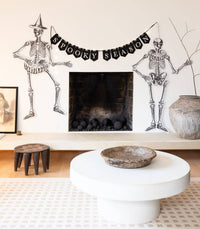 Living room with Halloween decorations including skeletons and a 'Spooky Season' banner hung across a mantel in a room, fairy lights illuminated.