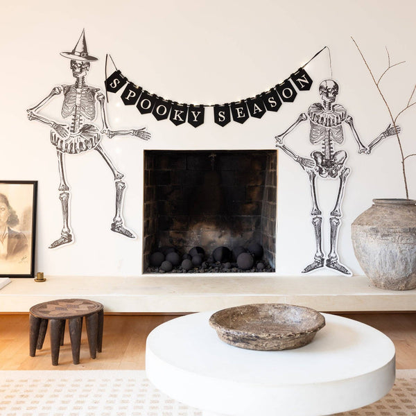 Living room with Halloween decorations including skeletons and a 'Spooky Season' banner hung across a mantel in a room, fairy lights illuminated.