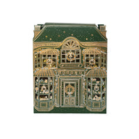 Front view of the green and gold foil paper toy store with bay windows, awnings, and wreaths, lit from inside