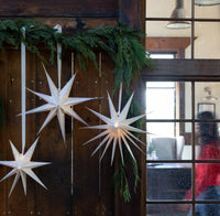 Two lit cream paper stars hanging on a rustic wood door with greenery.