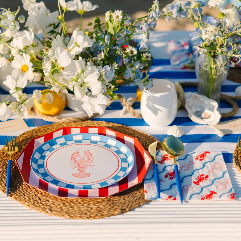 Overhead view of a seaside‑inspired place setting—a red‑and‑white striped octagon plate topped with a lobster plate, blue‑and‑white checked rim, and matching “lobster napkins,” all set on a woven charger with gold‑and‑blue flatware, against a bold blue striped runner, white wildflowers, lemons, and oyster shells scattered along the center.