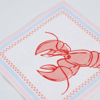 Angled close-up of a single paper cocktail napkin showing detail of the red line-drawn lobster illustration — claws extended, antennae visible, facing right. Periwinkle blue dotted inner border and pink outer edge clearly visible. Napkin is folded, square, white ground.