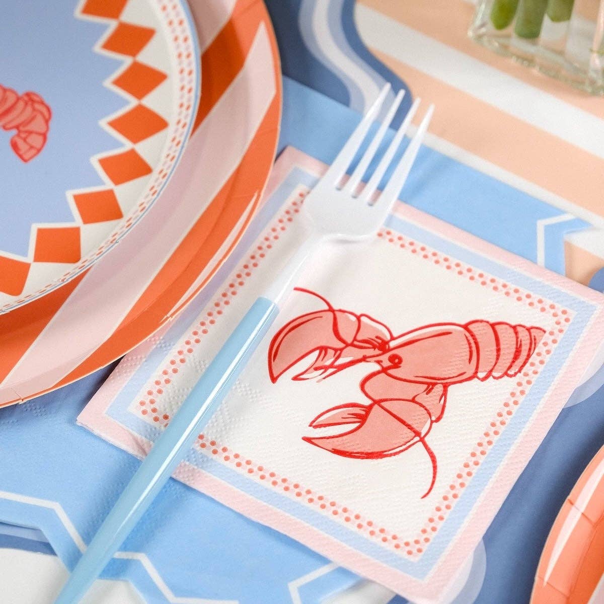 Close-up of a single lobster cocktail napkin placed to the right of a plate stack. Fork rests diagonally across the napkin. Periwinkle blue dotted border and pink outer edge in full detail. Plate border with orange diamond-dot pattern visible at left edge of frame.