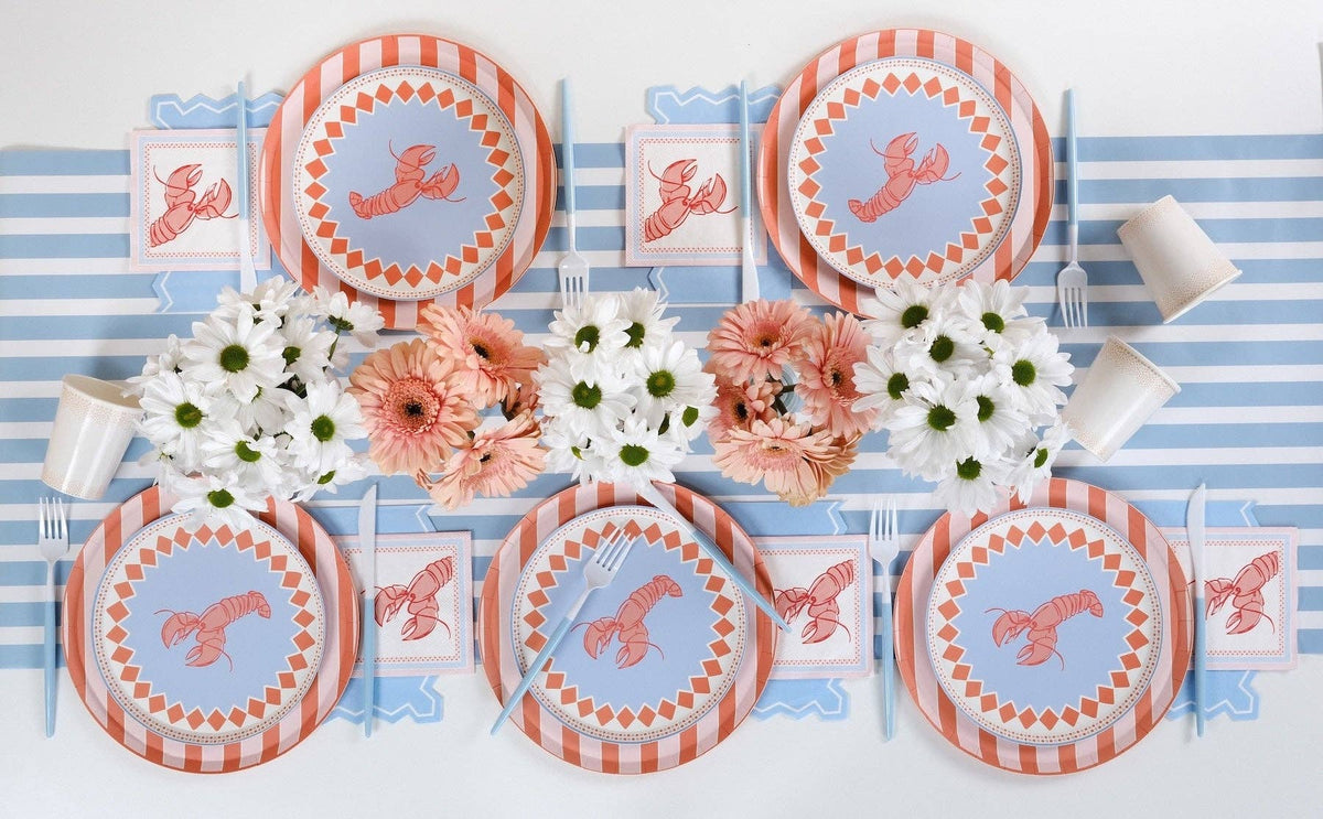 Overhead shot of a table set for six with a blue stripe table runner. Every place setting uses a large blue scalloped placemat under a coral-orange stripe scalloped charger plate topped with a periwinkle blue lobster dinner plate. Each place setting has a folded lobster cocktail napkin positioned to the left of the plate. Peach gerbera daisies and white daisies in bud vases centered.