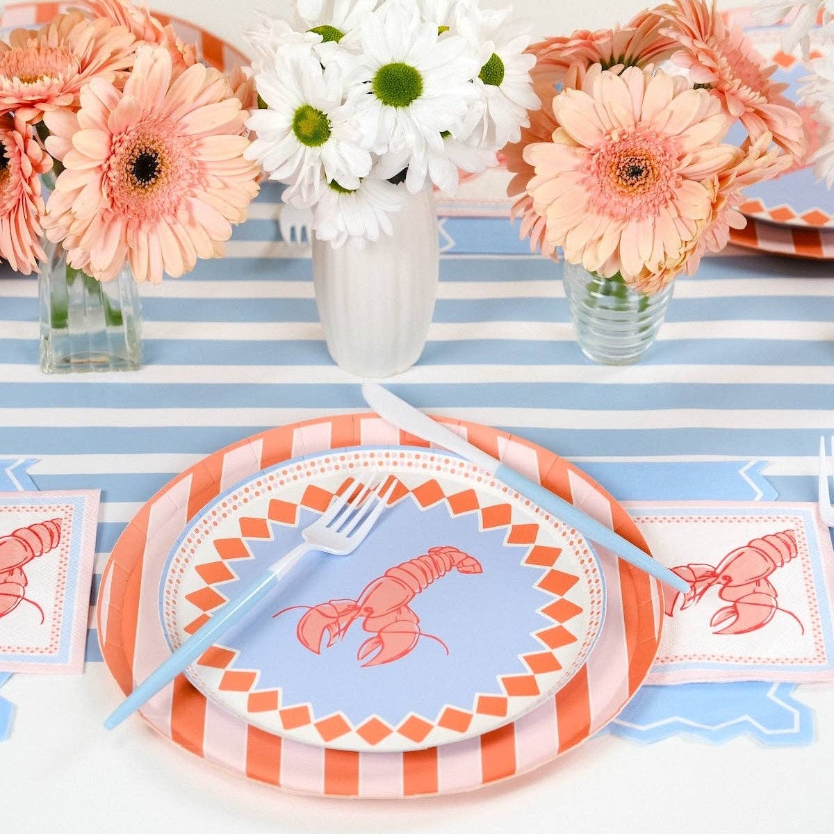 Angled view of a single place setting. Orange stripe scalloped dinner plate topped with periwinkle blue lobster print plate. Blue fork rests on the plate. Two lobster napkins visible flanking the setting on the blue stripe runner. Peach and white daisy arrangements in background.