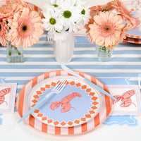 Angled view of a single place setting. Orange stripe scalloped dinner plate topped with periwinkle blue lobster print plate. Blue fork rests on the plate. Two lobster napkins visible flanking the setting on the blue stripe runner. Peach and white daisy arrangements in background.