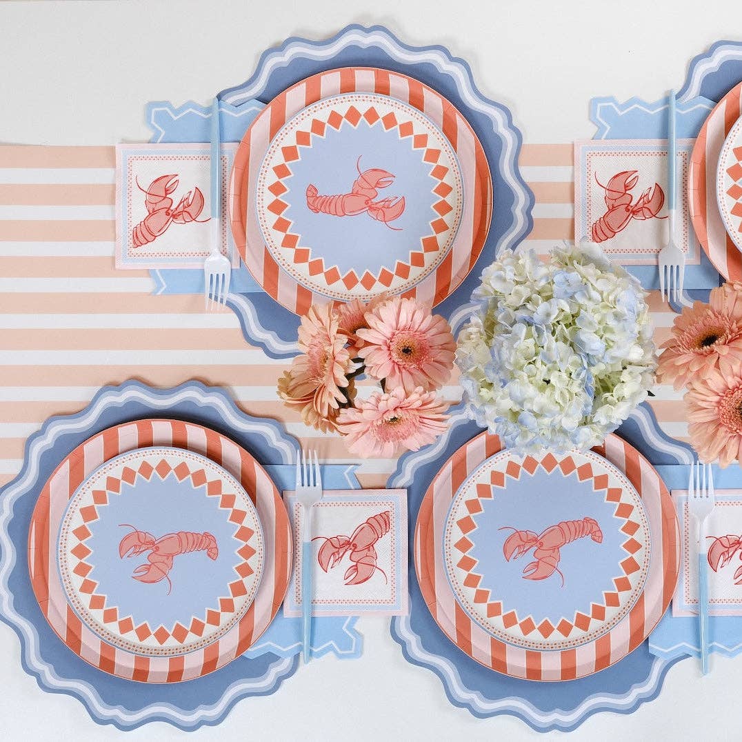 Overhead of two place settings on a peach and white diagonal stripe table runner. Blue scalloped placemat visible under each place setting. Lobster napkins folded square at each setting. Peach gerbera and blue hydrangea centerpiece. Blue scalloped table runner/border visible at table edge.