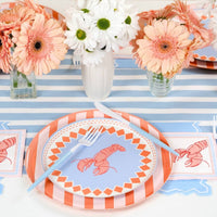 Overhead shot of a table set for four to six on a blue-and-white stripe table runner and white table surface. Each place setting has the lobster salad plate over the orange stripe charger, with a lobster cocktail napkin to the side. Blue plastic forks at every setting. White speckled paper cups at upper right. Peach gerbera daisies and white daisy bouquets as centerpieces.