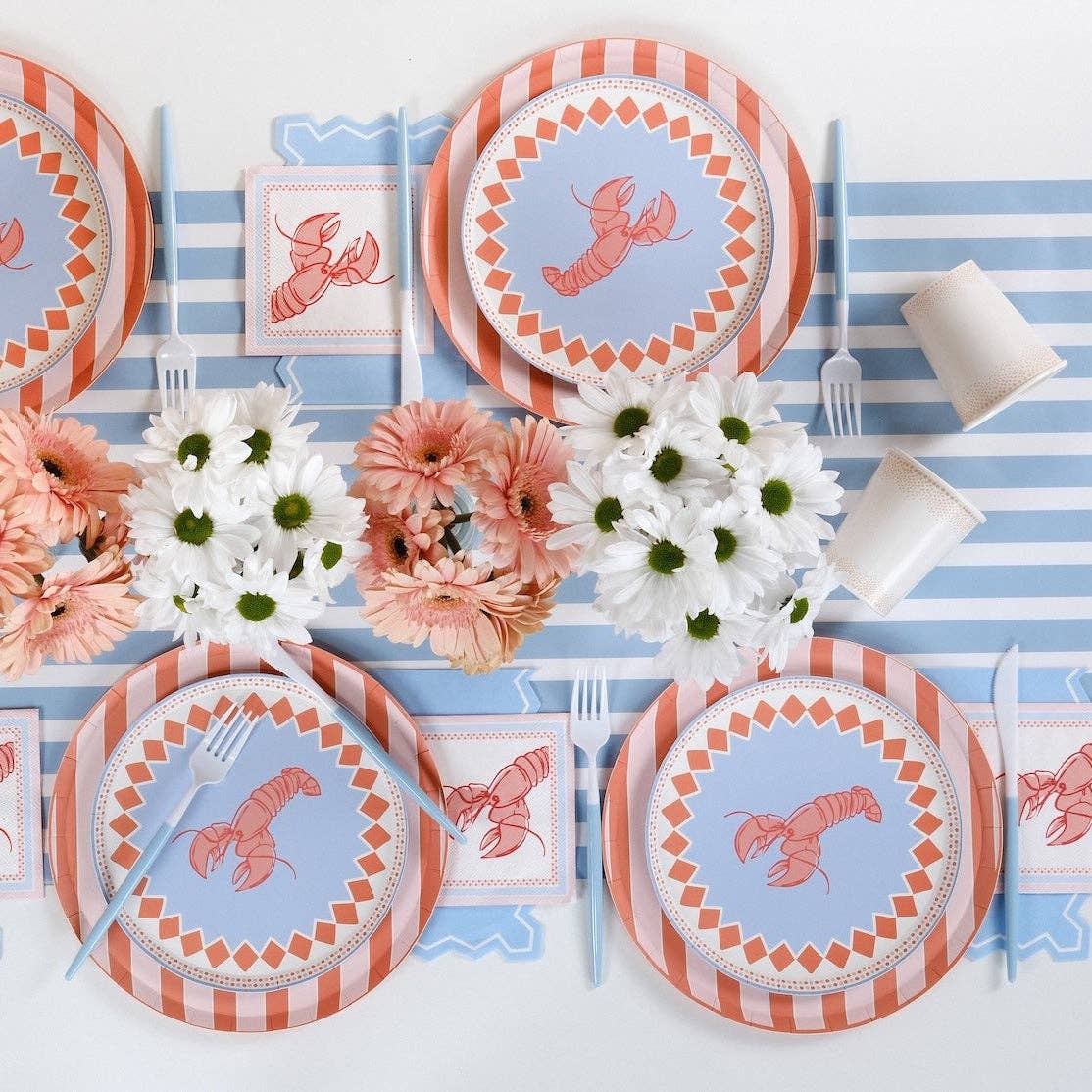 Wide overhead shot of a table set for four with a peach-and-white stripe table runner and large blue scalloped placemat/runner as the base layer. Each place setting layers the lobster salad plate over the orange stripe charger. Lobster napkins flanking each setting. Peach gerberas and blue-white hydrangea centerpiece. Blue plastic forks.